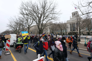Eine große Gruppe von Menschen nimmt an einer Protestdemo auf einer Straße in Washington, D.C. teil, einige halten Schilder und Transparente, andere fahren Fahrräder, und es gibt Schilder, Bäume und einen klaren blauen Himmel im Hintergrund.