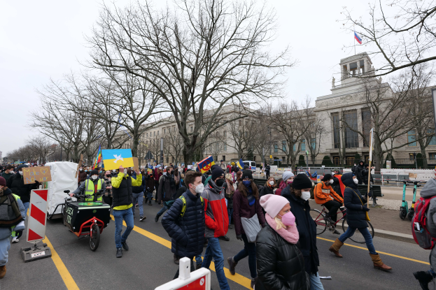 Eine große Gruppe von Menschen nimmt an einer Protestdemo auf einer Straße in Washington, D.C. teil, einige halten Schilder und Transparente, andere fahren Fahrräder, und es gibt Schilder, Bäume und einen klaren blauen Himmel im Hintergrund.