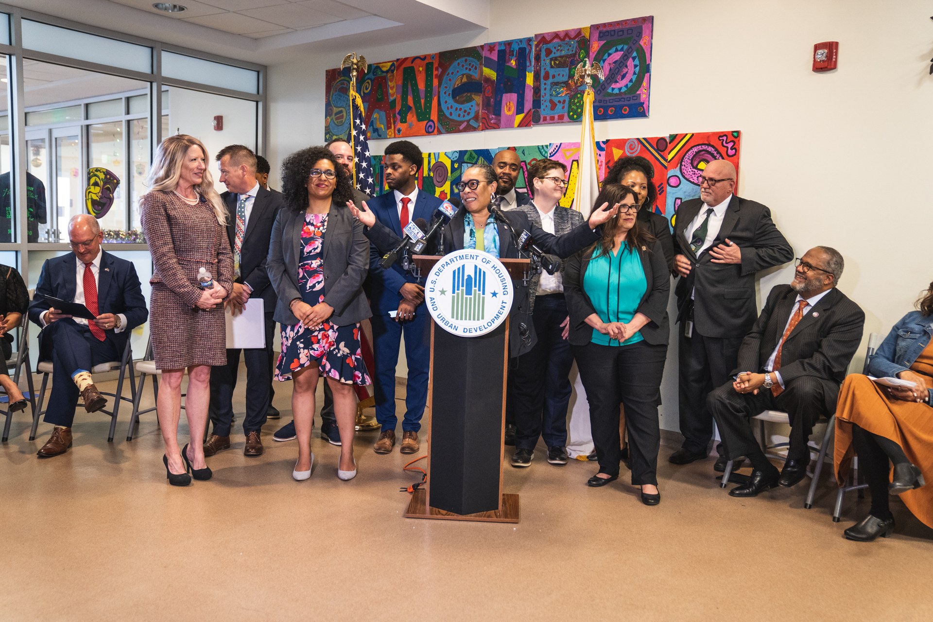 Eine Gruppe von Menschen steht vor einem Podium mit einem Mikrofon und einer Tafel mit dem Text "Sancho Stadtrat tritt dem Stadtrat bei", während andere auf Stühlen sitzen oder in der Nähe stehen; Flaggen, eine Tafel, ein Feuerlöscher und Deckenleuchten sind an der Rückwand zu sehen.