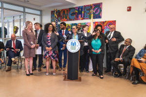 Eine Gruppe von Menschen steht vor einem Podium mit einem Mikrofon und einer Tafel mit dem Text "Sancho Stadtrat tritt dem Stadtrat bei", während andere auf Stühlen sitzen oder in der Nähe stehen; Flaggen, eine Tafel, ein Feuerlöscher und Deckenleuchten sind an der Rückwand zu sehen.