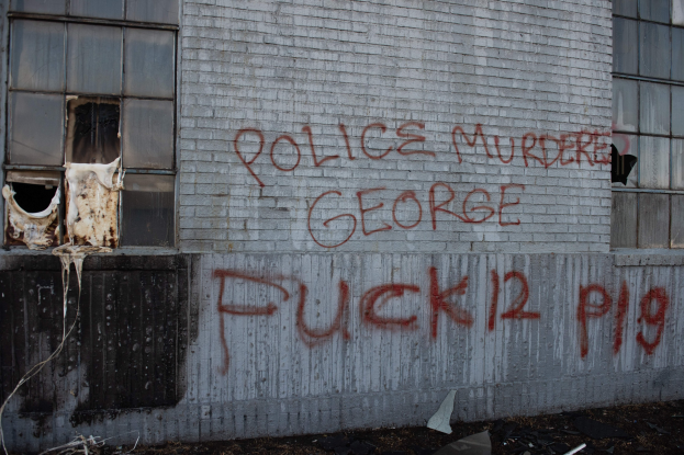 Graffiti an einer Gebäudewand mit der Aufschrift 'Polizeimord George Fuck 12 P19' und Fenstern sowie verstreuten Gegenständen davor.