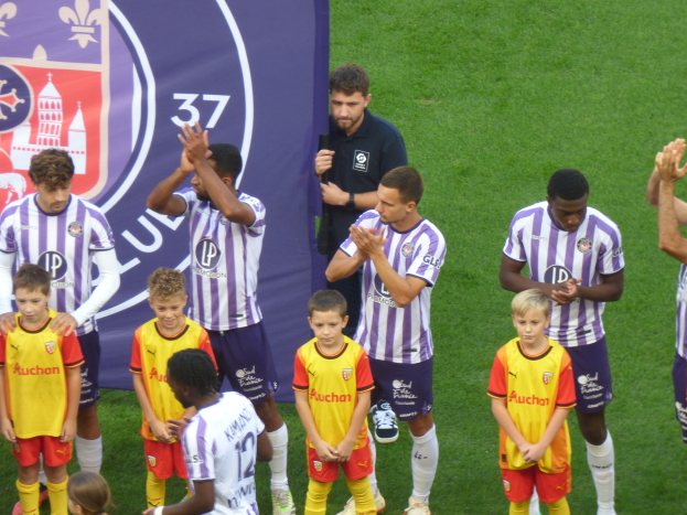 Eine Gruppe junger Jungs steht auf einem grünen Fußballfeld und klatscht in die Hände, mit einem Banner und Logos im Hintergrund.