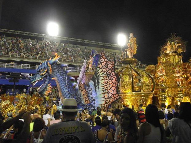 Eine große Menschenmenge um einen hell erleuchteten, farbenfrohen Festwagen an einem Karneval, mit einem Geländer um den Festwagen und einem Gebäude im Hintergrund unter einem dunklen Himmel.
