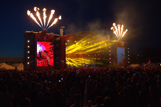 Eine große Menschenmenge auf einem Musikfestival schaut auf einer hell erleuchteten Bühne performende Künstler, mit Zelten links und Feuerwerk im dunklen Himmel dahinter.