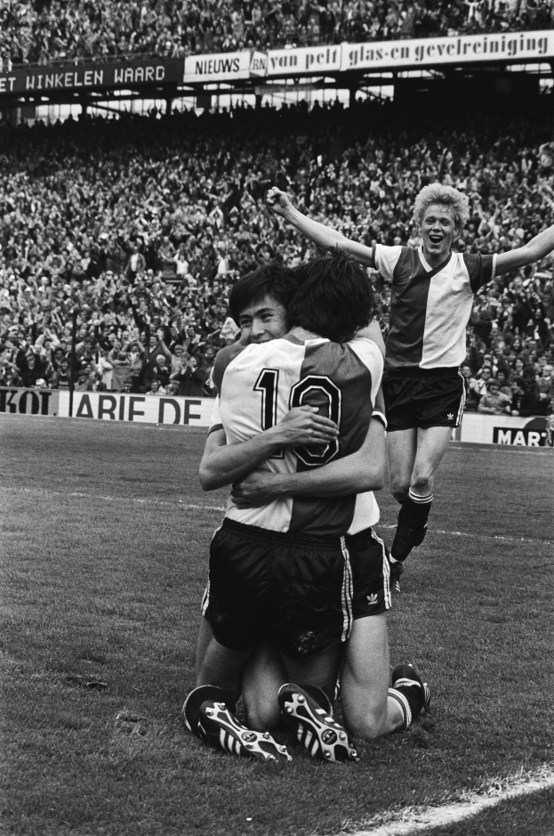 Drei Fußballspieler feiern auf dem Boden nach einem Tor, mit jubelnder Menge und schwenkenden Fahnen im Hintergrund, auf einem Schwarz-Weiß-Bild.