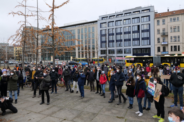 Große Gruppe von Menschen vor einem Gebäude mit Masken tragenden und Schilder haltenden Personen, Fahrzeugen, Bäumen und Gebäuden im Hintergrund bei einer Demonstration in Berlin.