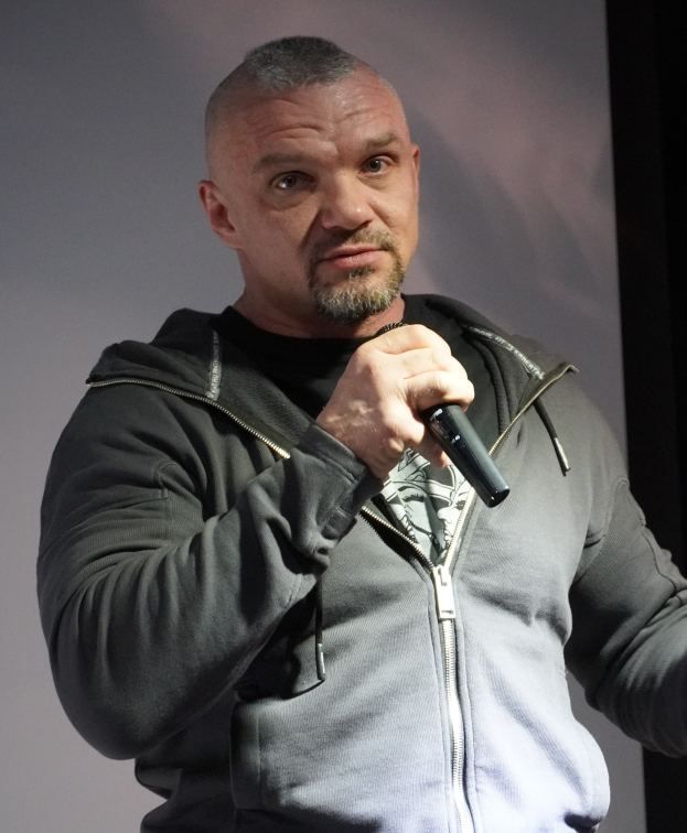 Comedian in einem schwarzen Anzug und weißem Hemd mit Mikrofon, selbstbewusst vor einer Wand stehend