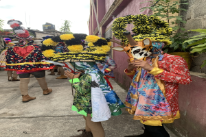 Eine Gruppe von Menschen in bunten Kostümen steht zusammen vor einem Gebäude im Freien und hält verschiedene Gegenstände, mit Bäumen, Gebäuden und einem klaren blauen Himmel im Hintergrund.