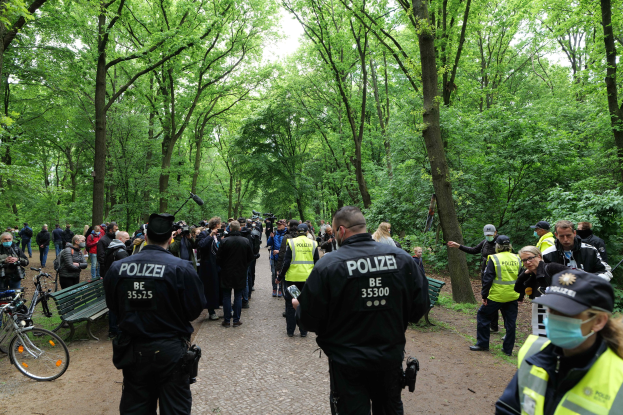 Polizeibeamte vor einer Menge während einer Outdoor-Veranstaltung, mit Fahrrädern und einer Bank im Vordergrund und Bäumen im Hintergrund.