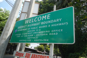 Ein grünes Schild mit der Aufschrift 'Willkommen an der Landkreisgrenze' ist an zwei weißen Pfählen befestigt, im Hintergrund sind Fahrzeuge, Gebäude und Bäume sowie ein blauer und weißer Himmel zu sehen.