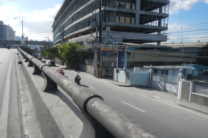 Eine Stadtstraße mit Fahrzeugen, einer großen Rohrleitung entlang des Straßenrandes, Gebäuden, Bäumen, Polen, Straßenlaternen, Strommasten mit Drähten und einem bewölkten Himmel.