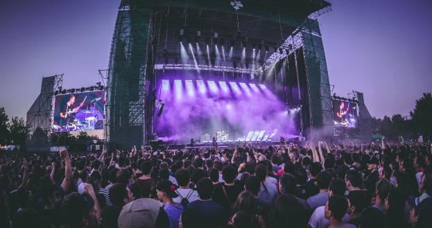 Eine große Menge Menschen bei einem Musikfestival schaut auf einer beleuchteten Bühne mit Instrumenten und Bildschirmen, die vor einem Gebäude, Bäumen und einem blauen Himmel steht.