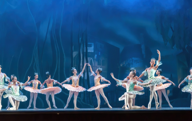 Eine Gruppe von Balletttänzern in farbenfrohen Kostümen, die *Der Nussknacker* auf der Bühne des Royal Opera House tanzen, bewegen sich synchronisiert gegen einen blauen Hintergrund.
