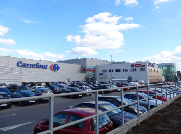 Großes Autohaus mit zahlreichen Fahrzeugen auf dem Hof, umgeben von Gebäuden, Straßeninfrastruktur und Fahrzeugen auf der Straße unter einem bewölkten Himmel.
