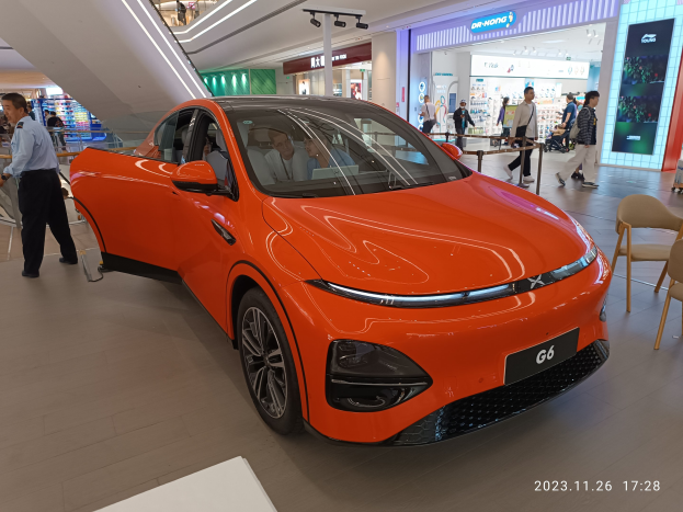 Orangefarbener Elektroauto in einer Ausstellungsfläche umgeben von Menschen, Stühlen, einem Wagen, Geschäften und Namensschildern im Hintergrund.