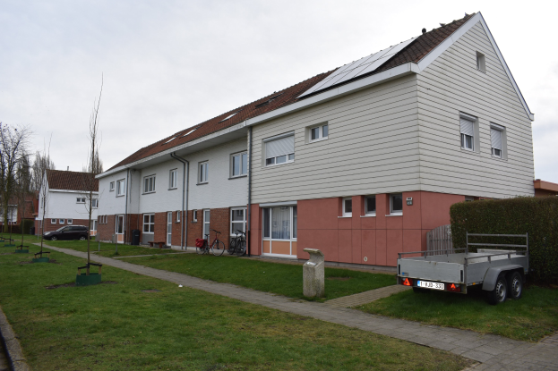 Ein Lastwagen vor einer Reihe von Häusern mit Solarpaneelen auf den Dächern, mit Fenstern, Türen und in der Nähe geparkten Fahrrädern, auf einer grasbewachsenen Fläche mit einem Weg, Pflanzen, Bäumen und einem bewölkten Himmel.