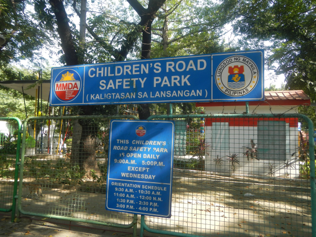 Metallzaun um einen Kindersicherheitspark mit zwei blauen Informationsschildern, einem kleinen Haus, Pflanzen, Bäumen und einem bewölktem Himmel.