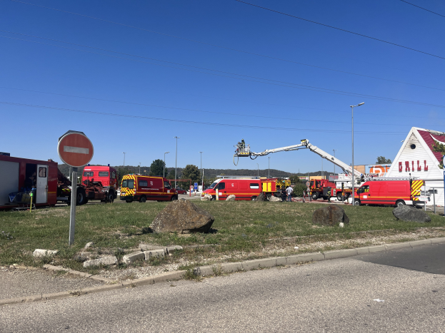 Gruppe von Feuerwehrautos auf einem grasigen Straßenrand neben einem Haus, Pfählen, Bäumen und einem bewölkten Himmel, wahrscheinlich nach einem Tankstellenbrand.