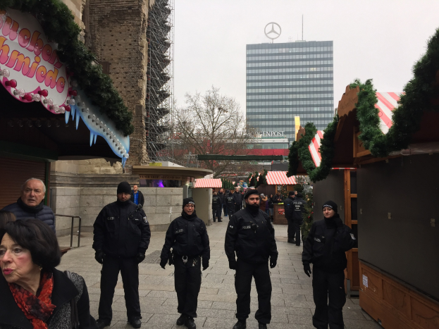 Eine Gruppe von Polizisten in schwarzen Uniformen und Mützen steht vor einem geschmückten Weihnachtsmarkt in Berlin, mit Gebäuden, Bäumen und einem klaren blauen Himmel im Hintergrund.