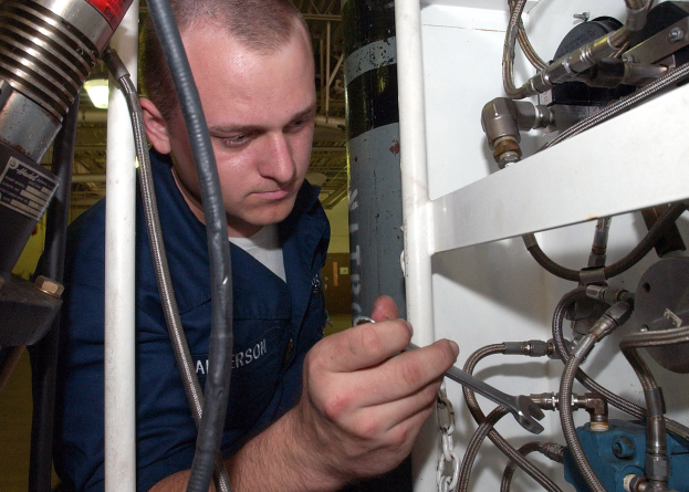 Techniker in blauer Uniform an einer Maschine mit Werkzeug, umgeben von Industrieausrüstung, Rohren und Beleuchtung.
