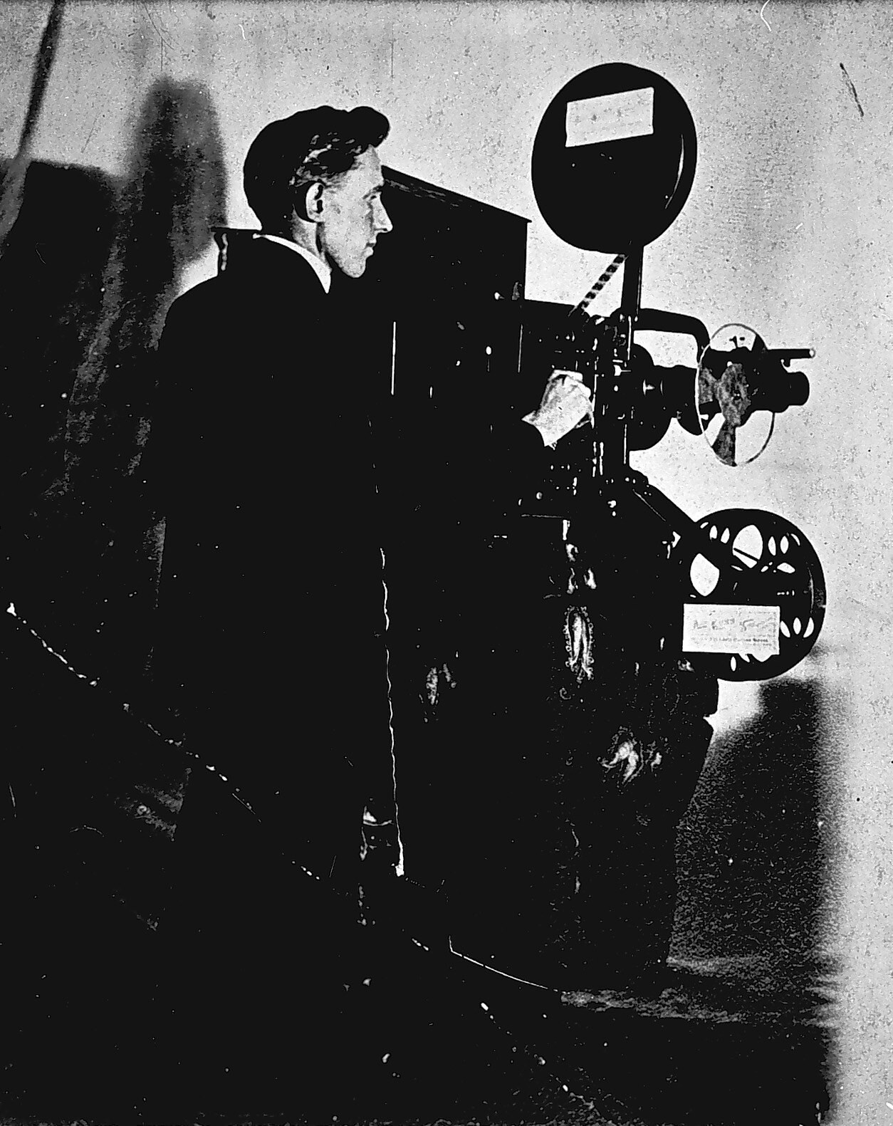 Altes Schwarz-Weiß-Foto eines Mannes in einem Anzug, der neben einem Filmprojektor steht und einen ernsten Gesichtsausdruck hat.