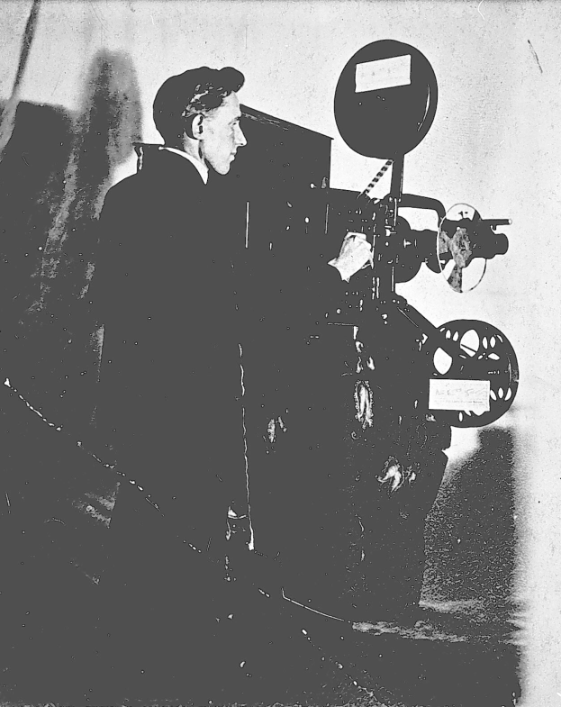 Altes Schwarz-Weiß-Foto eines Mannes in einem Anzug, der neben einem Filmprojektor steht und einen ernsten Gesichtsausdruck hat.