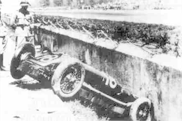 Ein Schwarz-Weiß-Foto eines verunglückten Rennwagens mit zwei Personen links daneben und Bäumen im Hintergrund.