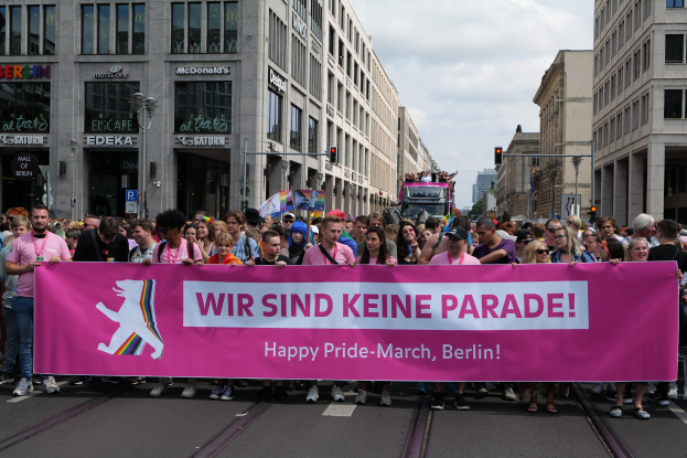 Gruppe von Menschen, die auf einer Straße in Berlin, Deutschland, mit einer pinken Fahne mit der Aufschrift "Happy Pride March" marschieren, mit Gebäuden, Laternenmasten und Verkehrszeichen an der Straße unter einem bewölkten Himmel.