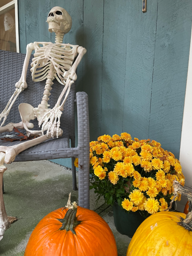 Ein Skelett sitzt auf einem Stuhl neben zwei Kürbissen auf einer Veranda, mit einem Blumentopf voller gelber Blumen in der Nähe und einer Wand mit einem Glasfenster im Hintergrund.