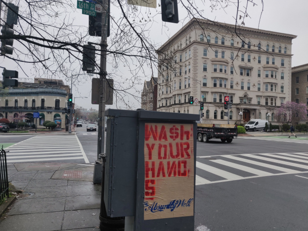 Eine belebte Stadtstraße mit Fahrzeugen, einem Fußgänger, einem "Waschen Sie Ihre Hände"-Müllcontainer, Verkehrsampeln, Schildern, einem Metallzaun, Pflanzen, Bäumen, Gebäuden und einem klaren blauen Himmel.