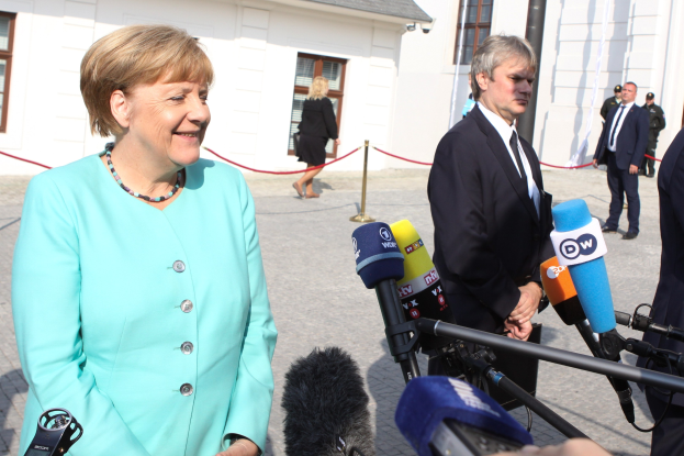 Bundeskanzlerin Angela Merkel beim Sprechen mit Reportern vor einem Gebäude mit einer Gruppe von Menschen, die Mikrofone und eine Absperrung halten, bei bewölktem Himmel.