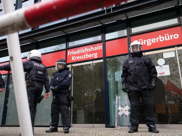 Eine Gruppe von Polizisten in Helmen und Handschuhen steht vor einem gläsernen Gebäude, mit einem Pfahl links und Schildern im Hintergrund, die "Friedrichshain Kreuzberg" lesen, und Papieren an den Glaswänden geklebt.