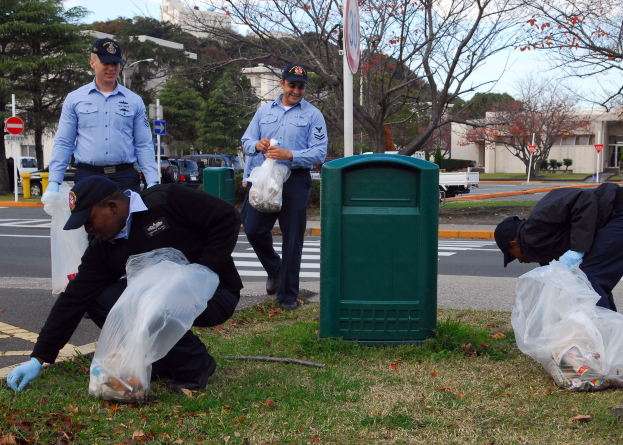 Drei Polizisten in Uniform sammeln Müll an einer Straße, mit Bäumen, Gebäuden, Fahrzeugen und einem Mülleimer im Hintergrund.