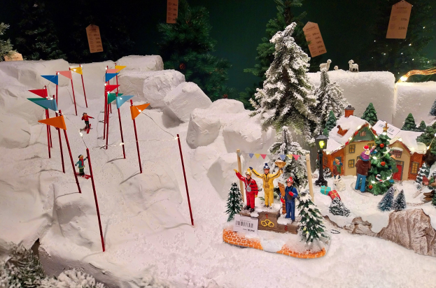 Eine festliche Weihnachtsdorfszene mit schneebedeckter Berglandschaft im Hintergrund, mit verschiedenen geschmützten Objekten und Figuren unter einer dunklen, gemütlichen Atmosphäre.