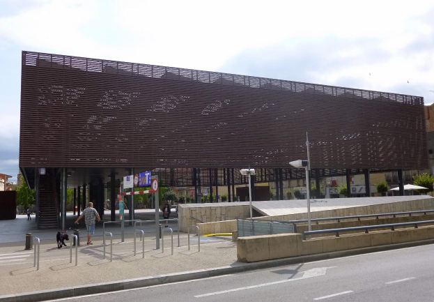 Großes Gebäude mit zahlreichen Fenstern, das als neuer Eingang zur Stadtmitte dient, flankiert von Straßeninfrastruktur, Fahrzeugen, Fußgängern und Bäumen unter einem bewölkten Himmel.