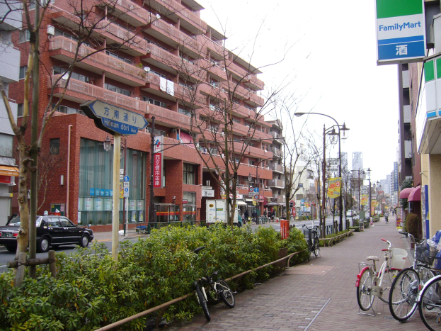 Eine Stadtstraße mit parkenden Fahrrädern, Gebäuden, Straßenlaternen, Fahrzeugen, Fußgängern, Grünflächen und einem klaren Himmel.