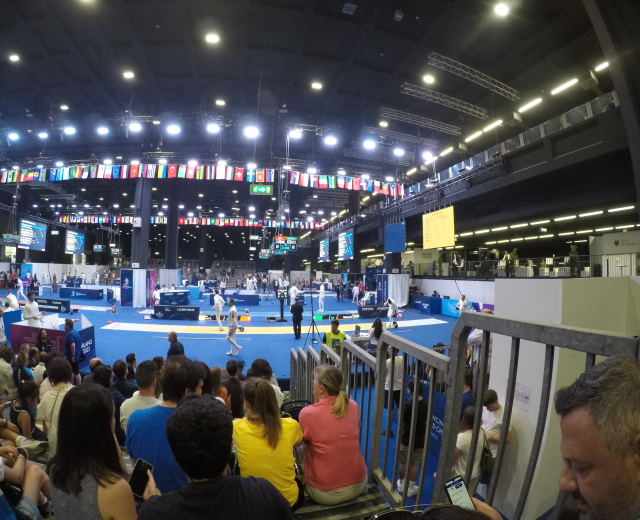Große Menschenmenge sitzt in einer Indoor-Sportarena und schaut auf eine Bühne, mit Stehplätzen, Bildschirmen, Fahnen und Deckenleuchten im Hintergrund.