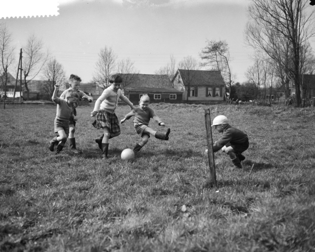Eine Gruppe von Kindern, die Fußball auf einem Feld spielen, mit einem Ball in der Mitte der Aktion, umgeben von Bäumen und Häusern, unter einem sichtbaren Himmel, dargestellt in Schwarz-Weiß.