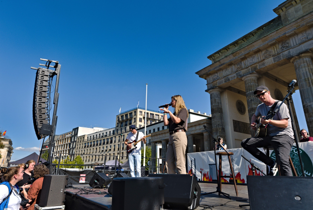 Eine Gruppe von Menschen, die auf einer Bühne vor dem Brandenburger Tor in Berlin, Deutschland, mit Musikinstrumenten, Mikrofonen, Lautsprechern und verstreuten Gegenständen spielt, vor einem Hintergrund aus Gebäuden, Bäumen, Pfählen, Fahnen und einem klaren blauen Himmel.