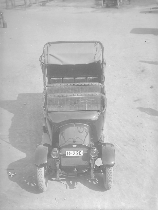 Altes Schwarz-Weiß-Foto eines Autos auf einem Parkplatz mit sichtlicher Nummernschild, andere Fahrzeuge im Hintergrund.