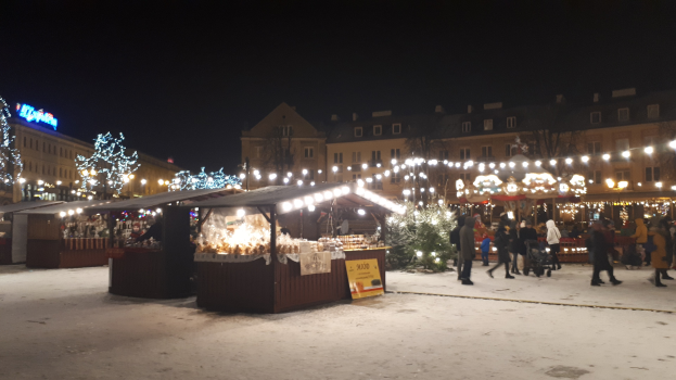 Ein verschneiter nächtlicher Weihnachtsmarkt mit Menschen, beleuchteten Ständen, Pflanzen, Bäumen, Gebäuden und Schildern unter einem bewölkten Himmel.
