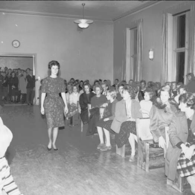 Eine Frau in einem schwarzen und weißen Kleid läuft über einen Laufsteg während einer Modenschau für Frauenrechte, mit einer Menge, die auf Stühlen sitzt und einer Uhr und Lichtern an der Wand.
