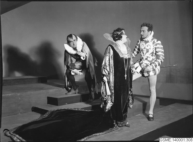 Schwarzes und weißes Foto von drei Menschen auf einer Bühne, wobei einer ein Kostüm trägt und einen Stock hält, ein anderer auf einer Treppe sitzt und unten ein Text mit "Der Phantom der Oper" steht.