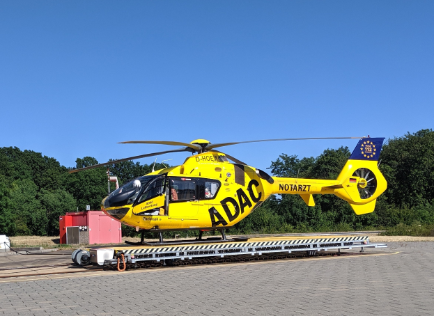 Ein gelber Hubschrauber steht auf dem Rollfeld eines Flughafens, umgeben von Bäumen und einem klaren blauen Himmel, mit verschiedenen Gegenständen rund um den Hubschrauber auf dem Rollfeld.