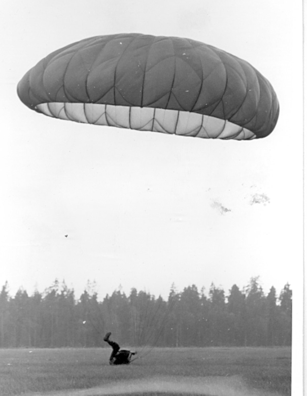Schwarzes und weißes Foto einer Person beim Paragleiten in der Luft mit einem Fallschirm, umgeben von Bäumen und einem klaren Himmel, mit Text unten.