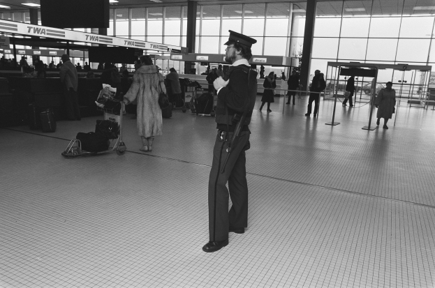 Ein Polizist in Uniform steht in einem Flughafen-Terminal, hält eine Waffe und ist von Menschen und Gepäck umgeben, mit Gepäckwagen, Deckenleuchten, Pfosten, Glaswänden und Informationsanzeigen im Hintergrund.