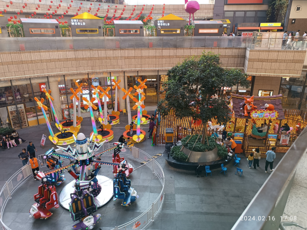 Ein Vergnügungspark innerhalb eines Einkaufszentrums mit verschiedenen Attraktionen, Bäumen, Menschen und Ständen, umgeben von Gebäuden, mit einem Wasserzeichen in der rechten unteren Ecke.