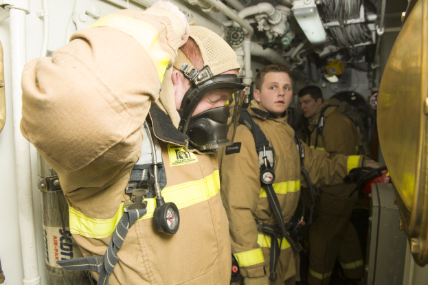 Feuerwehrleute in Gasmaske und Schutzausru├čtung stehen in einem Raum mit Rohren und Gegenst├Ąnden, mit einem Metallgegenstand auf der rechten Seite.