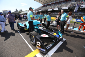 Eine Gruppe von Menschen, die um ein Formula-E-Rennwagen auf der Strecke feiern, mit einem Stadion im Hintergrund, das von Zuschauern unter einem klaren blauen Himmel gefüllt ist.