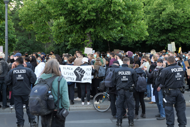 Eine große Gruppe von Menschen steht an der Straße, einige halten Schilder und tragen Mützen und Taschen, vorne ein Fahrrad und im Hintergrund Bäume und ein Pfahl während einer Black Lives Matter Demonstration in Berlin.